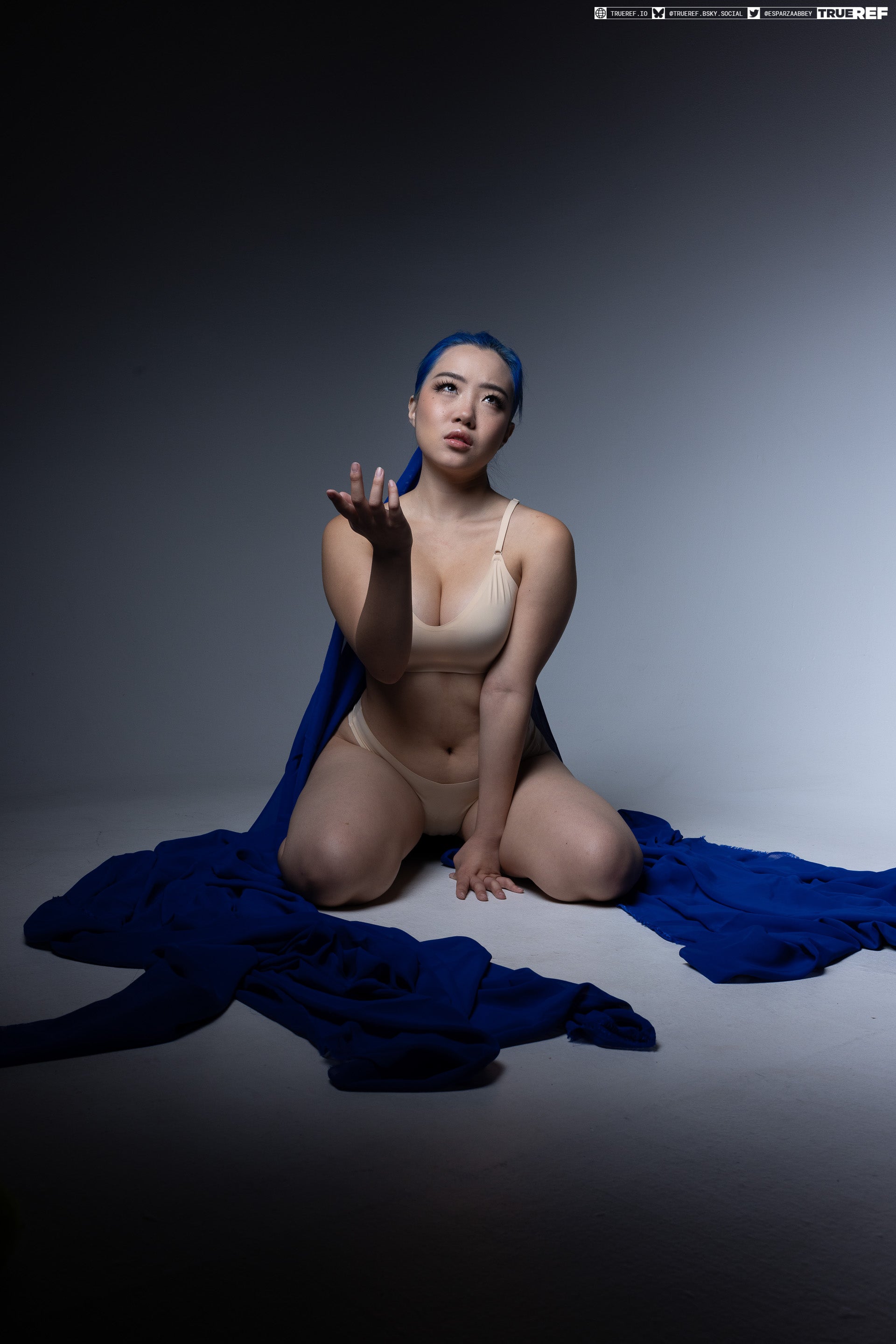 a studio shot of a woman kneeling with her hand outstretched and looking up, draped with a blue cloth and wearing underwear, with blue hair.