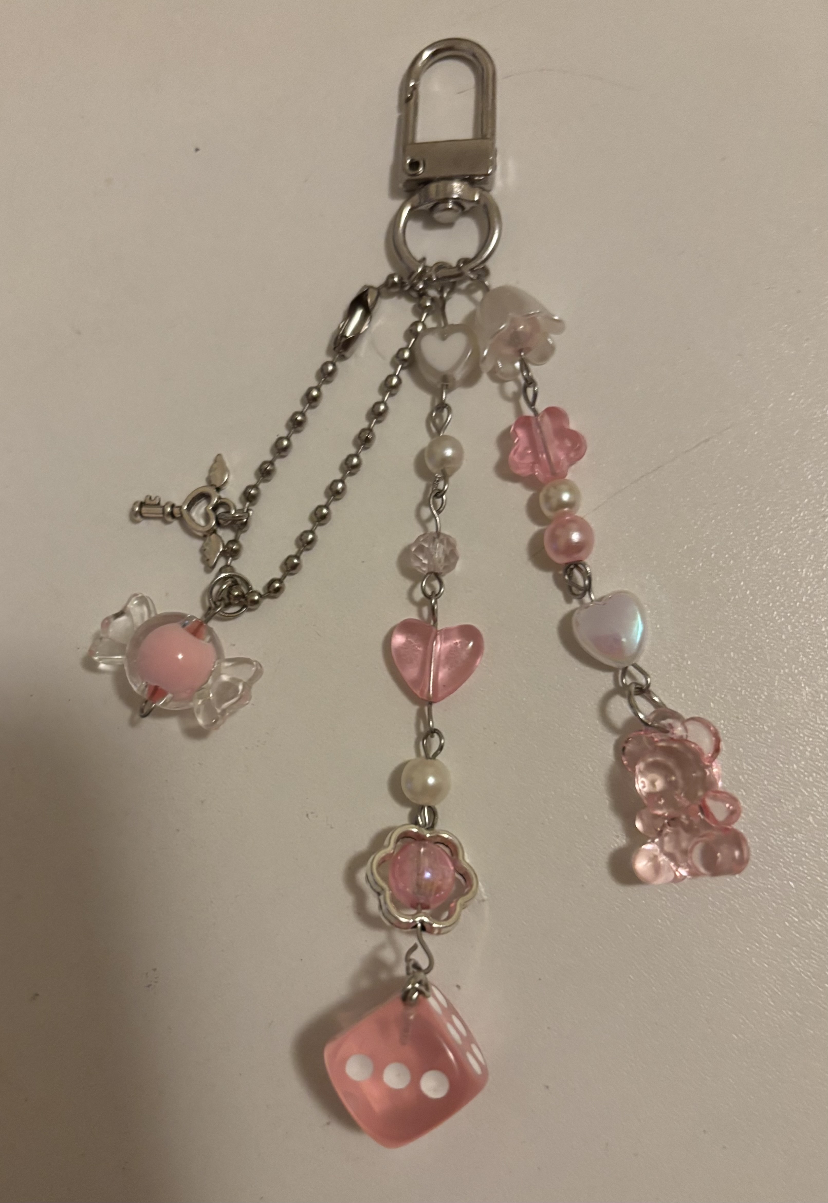 a closeup shot of a keychain featuring various pink and silver charms and beads. the charms include a candy, hearts, flowers, a die, and a bear. the beads are strung together with silver chains and are mostly pink and pearlcolored. the keychain has a silver clasp at the top.