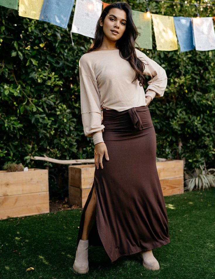 a woman is standing on a lawn, posing for the camera. she is wearing a long, brown, tiewaist skirt with side slits and a light beige longsleeved shirt.  she is also wearing light beige suede ankle boots.  behind her, there is a wooden planter box and a hedge.  above her, strung between trees, are colorful pieces of fabric.  the lighting is natural and the image is taken from eye level.