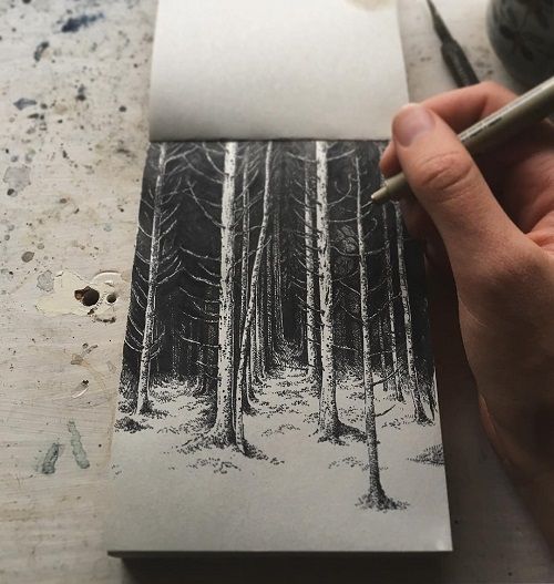 the image shows a closeup of an artist's hand meticulously drawing a detailed pen and ink illustration of a dark, dense forest scene in a sketchbook.  the artist is using a fineliner pen, and the drawing is highly detailed, employing techniques like stippling and hatching to create texture and depth in the depiction of the trees and the forest floor. the sketchbook lies on a textured, worn surface, possibly a table or workbench. the overall tone is dark and moody, reflecting the atmospheric nature of the forest illustration. the focus is on the drawing itself and the process of its creation.