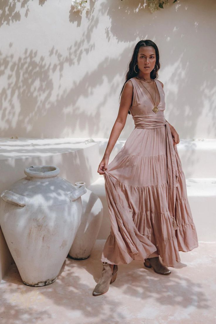a woman in a long, flowing, tiered, pink maxi dress stands outdoors against a white wall.  she is wearing gold jewelry, including a necklace and earrings, and brown ankle boots. the dress has a wrap style bodice and a layered skirt.  the setting appears to be a garden, with large white pots or vases nearby.  the sunlight casts shadows on the wall and the woman. the overall style is bohemian or bohochic.
