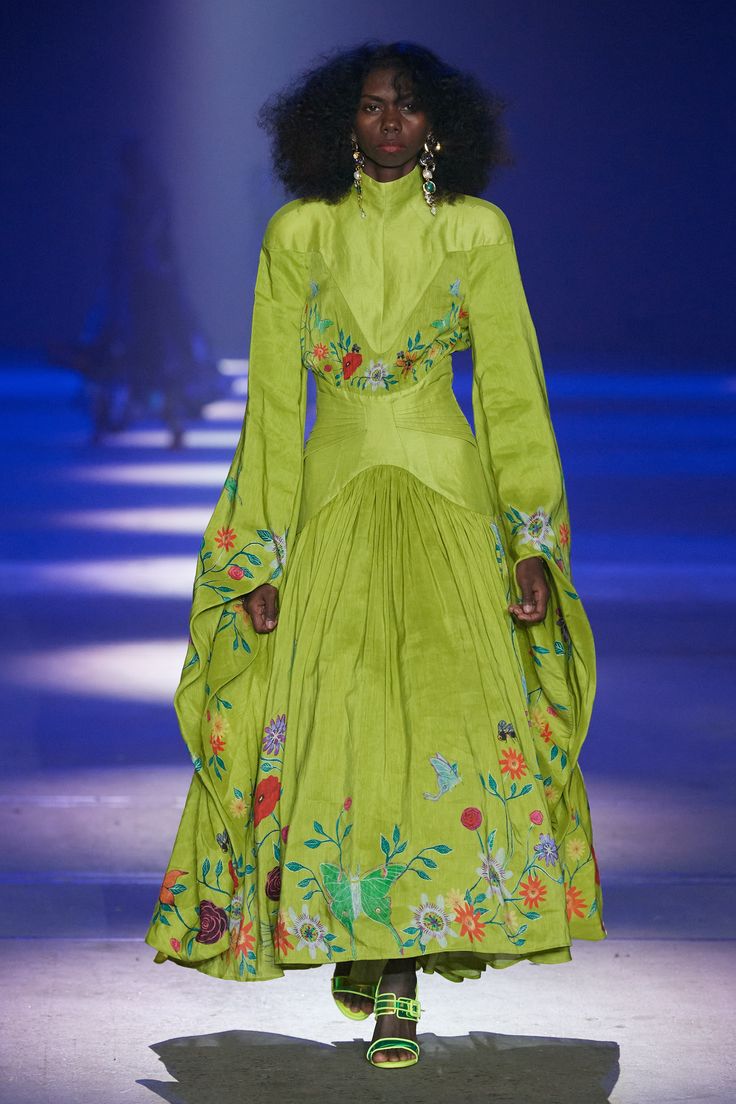 a black woman with afro hair walks down a runway in a lime green, anklelength dress with floral embroidery on the bodice and the bottom of the skirt. she wears large chandelier earrings and lime green sandals. there are other people and lights behind her.