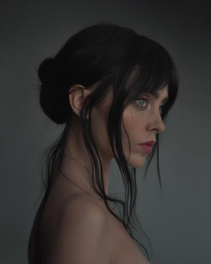 a portrait of a fairskinned woman in profile against a gray background. her dark hair is styled in a bun, with some strands falling loose around her face. she has blue eyes, red lips, and is wearing a delicate necklace. she appears to be looking away with a serious expression. her shoulder is bare. the overall aesthetic is artistic and ethereal.