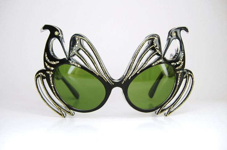 a closeup shot showcases a pair of unique sunglasses. the frames are black and shaped like stylized birds, adorned with embedded gemstones or beads. the lenses are a dark green tint. the sunglasses are placed on a white reflective surface.