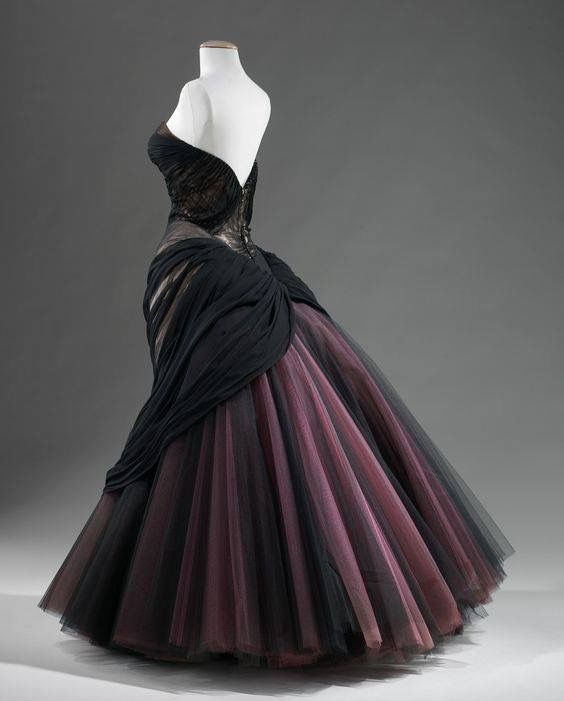 the image is a photograph of a strapless ball gown on a mannequin. the bodice is black lace and the skirt is black and purple tulle. a black swath of fabric drapes from the waist down the side of the gown.