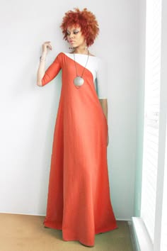 a woman with red hair is wearing a long orange dress with a white overlay and a silver pendant necklace. she is standing and looking down, and she has a serious expression on her face. the image is taken from a slightly low angle.  she is standing near a white wall with a window to the right. the image is welllit and there is no background clutter.