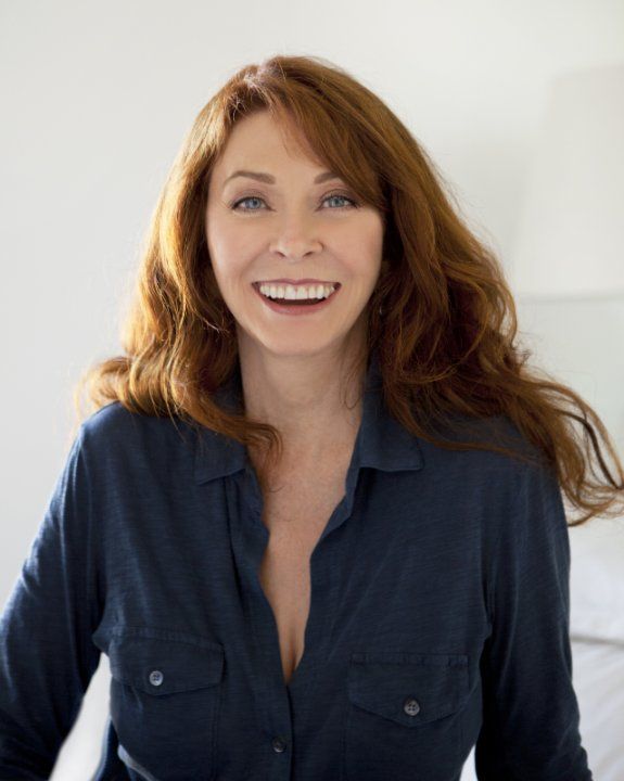 a smiling middleaged woman with long, red hair is wearing a blue buttondown shirt and looking directly at the camera. the background is white and the photo appears to be taken indoors.