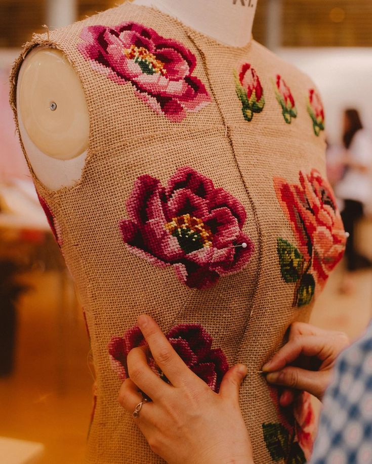 a closeup shot shows a dress form covered in burlap and adorned with crossstitch embroidery in a floral design, featuring peonies. hands are visible, pinning fabric to the dress form, suggesting the creation or alteration of a garment. the piece combines elements of traditional needlework with contemporary fashion design.