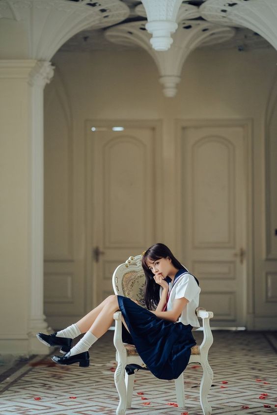 the image showcases a young asian woman sitting gracefully on a vintagestyle white chair. she is dressed in a classic school uniform consisting of a white shirt, a dark blue skirt, and black leather shoes. her long black hair cascades over her shoulders, framing her delicate features. she strikes a thoughtful pose, her hand resting lightly on her chin. the setting is a grand hallway with ornate white walls, arched doorways, and a tiled floor. soft natural light streams in from the doorways, illuminating the scene with a gentle and ethereal glow.