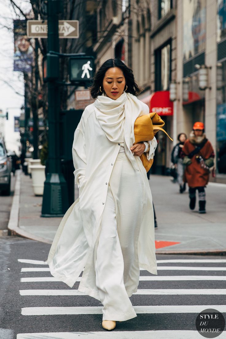 a woman in allwhite outfit, including a long coat and matching pants, walks across a crosswalk in a city street. she wears a large, draped scarf around her neck, and carries a yellow bag in her left hand. she's looking down as she walks. the background is a typical new york city street, with buildings, traffic lights, and pedestrians.