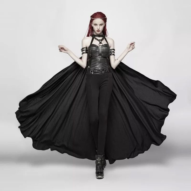 a full shot of a model wearing a complete gothic outfit. the outfit includes a long black cape, a black corset top with metal accents, and black pants. the model also wears black boots and has long red hair. the background is a plain grey. the overall style is dark, dramatic, and elegant, with elements of gothic, steampunk, and dark academia.