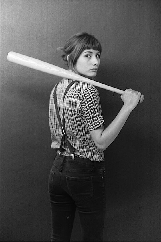 a black and white portrait of a young woman wearing a plaid shirt, jeans and suspenders. she is holding a baseball bat behind her and looking seriously at the camera. the image is shot in a studio setting.