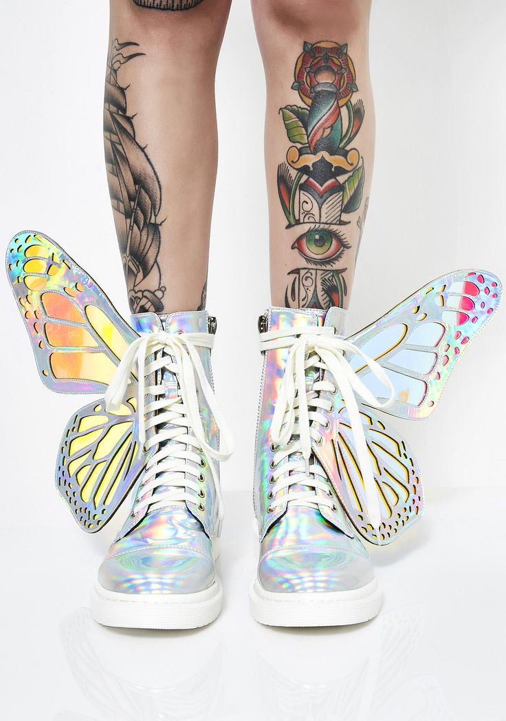 a person stands wearing holographic boots with butterfly wings attached to the back. the person has tattoos on their legs, including an eye, a dagger, and a rose. the boots have white laces and white soles. the background is white.