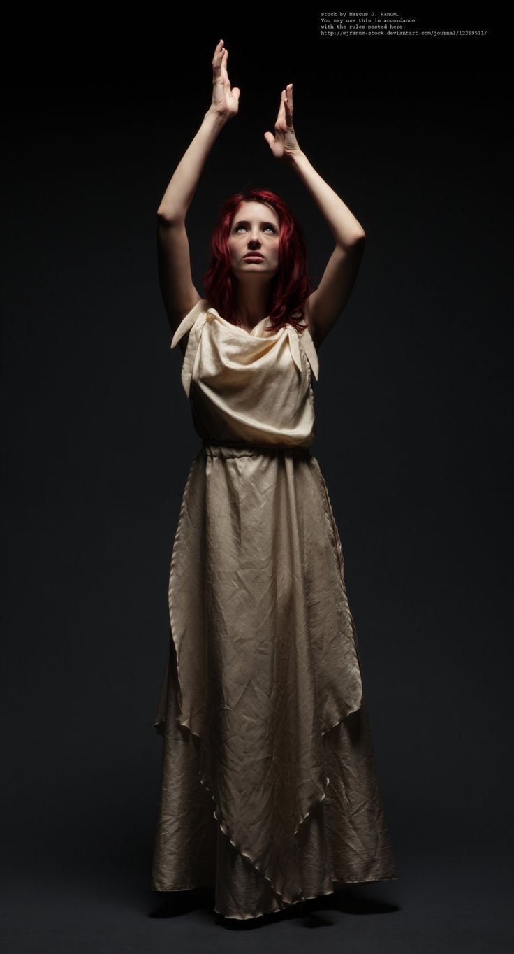 a woman with red hair wears a long white dress, she has her arms raised above her head and is looking up. she is standing in a studio on a dark background.