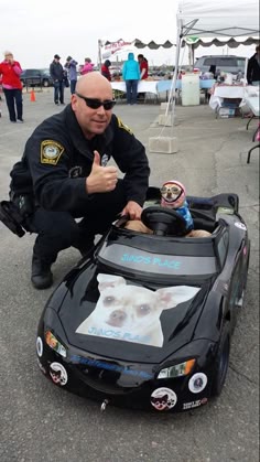 a police officer wearing a uniform and sunglasses is standing with his thumb up, in front of a black toy car, a chihuahua wearing a blue and white shirt is inside the car, driving it, with a happy expression.