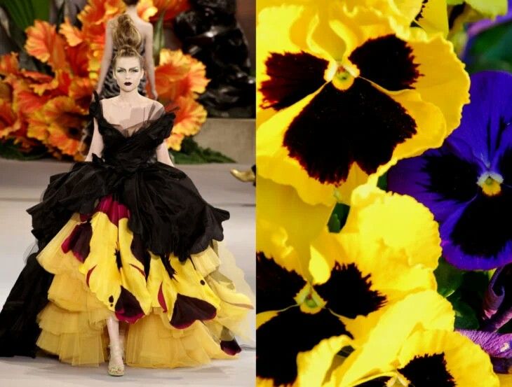 a fashion model is wearing a striking yellow and black dress with a floral pattern. the dress is very full and flowing, with multiple layers. the model is posing on a runway. in the background, there are many colorful pansies with yellow and purple petals.