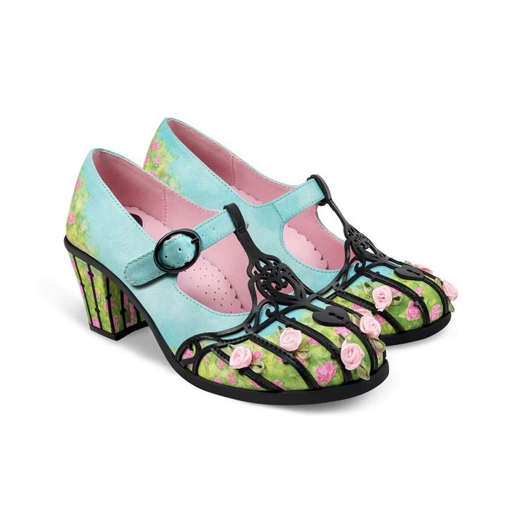 a pair of mary jane style dress shoes with a floral design on the front and heel are arranged on a white surface. the shoes have a light blue upper with a pink lining and a strap with a black buckle. the front of the shoes have a design resembling a flowerbed with pink roses and green foliage, enclosed by a black lattice structure. the heels feature a striped pattern in green and black, coordinating with the floral motif. the shoes are positioned closely together, with the front shoe slightly overlapping the back one.