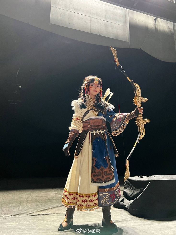 a woman in asian traditional clothing holds a bow and arrow on a stage. she has braids in her hair and is wearing fur and jewelry. she is wearing brown footwear.