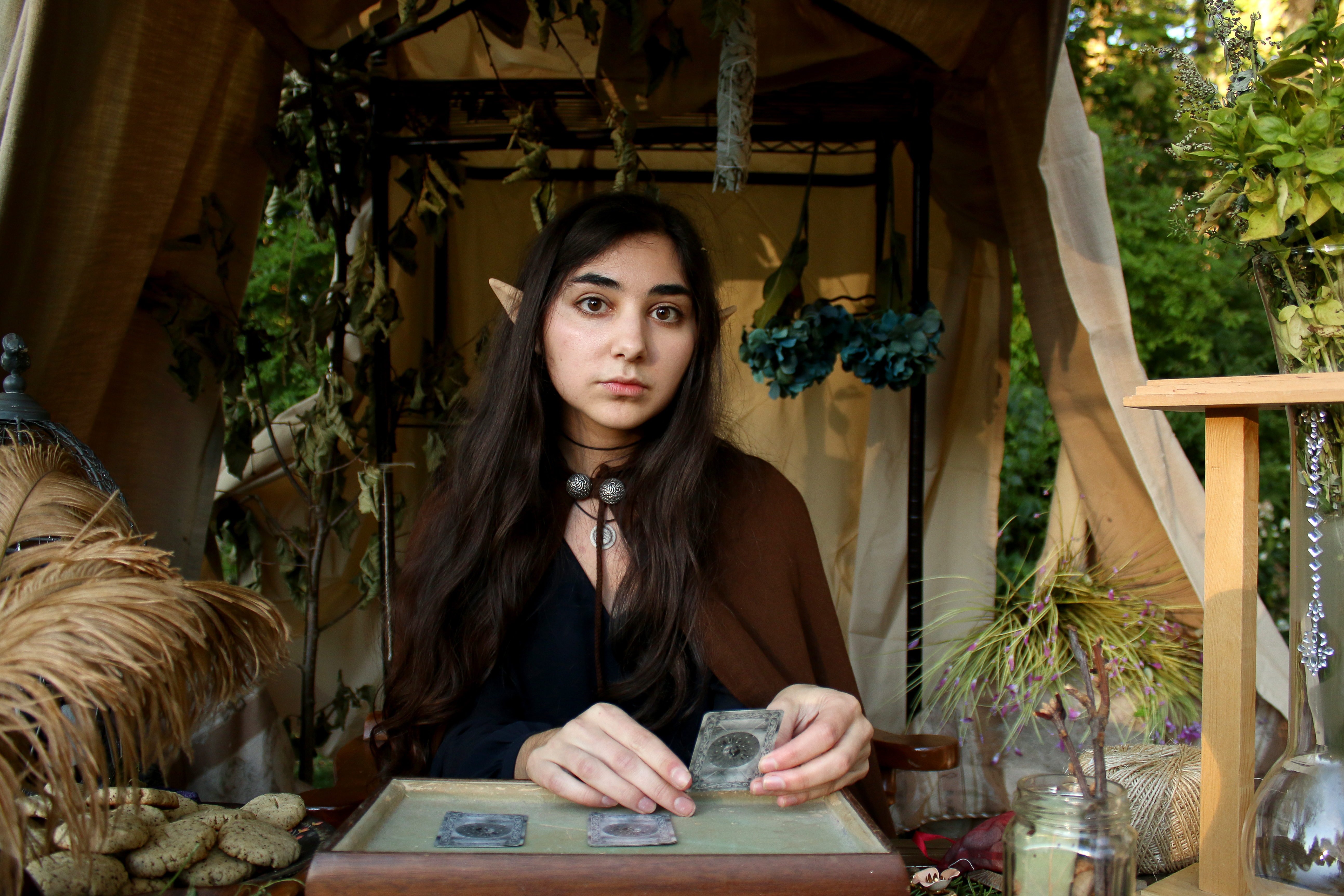 a young woman with elflike ears sits at a stall, seemingly reading tarot cards.  she's dressed in a dark, cloaklike garment and wears several necklaces. the stall is decorated with various herbs, plants, crystals, and other mystical items, creating an enchanting and atmospheric scene reminiscent of a fall or autumn market or fair. the overall ambiance is mystical and spiritual, suggesting a setting for fortunetelling or a magical practice.  she has a serious and focused expression.