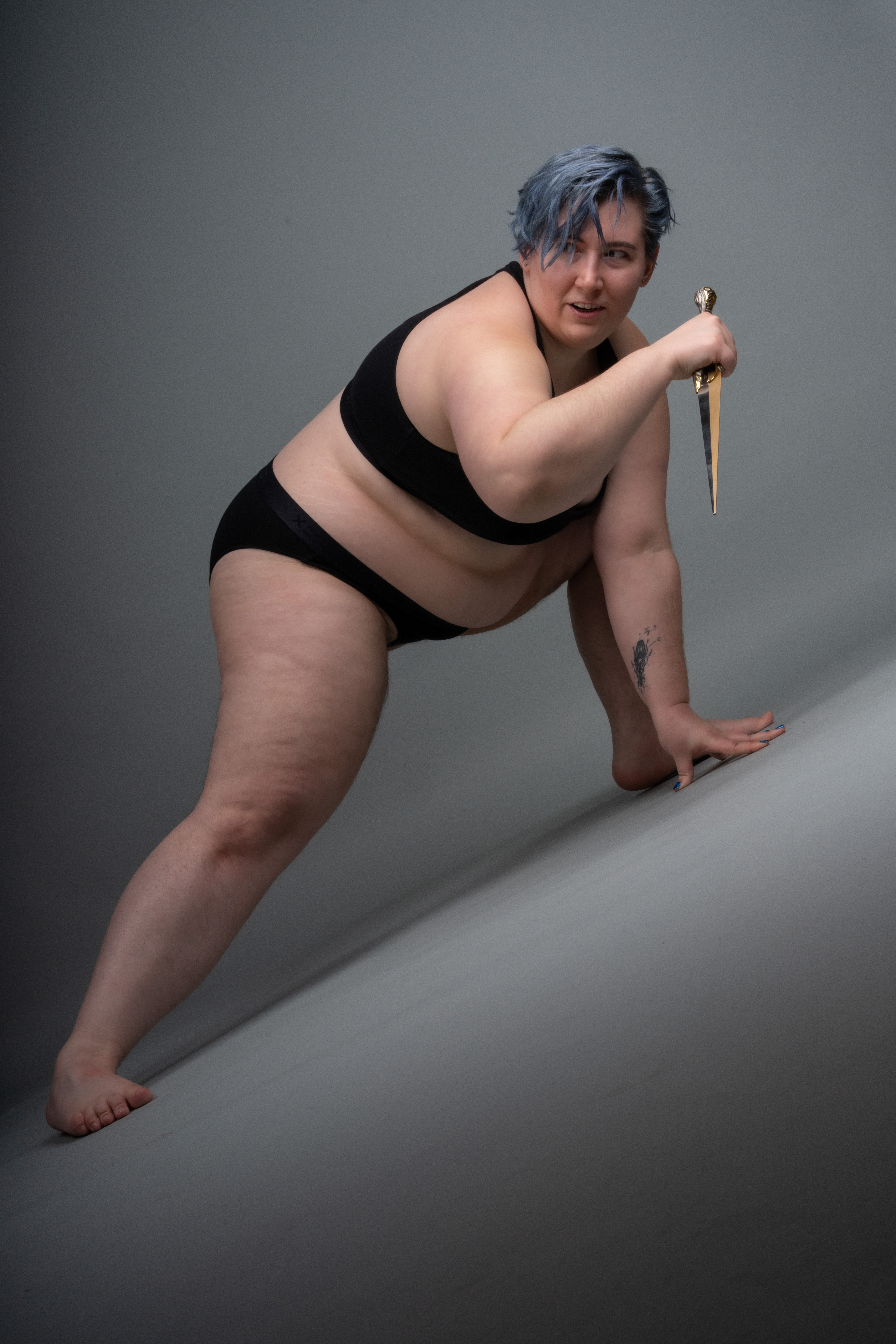 a full shot of a plus size woman with blue hair crouching and holding a dagger. she is barefoot, wearing black underwear and a black sports bra. she has a tattoo on her arm and is looking at the camera with an aggressive expression. the background is a plain grey studio backdrop.