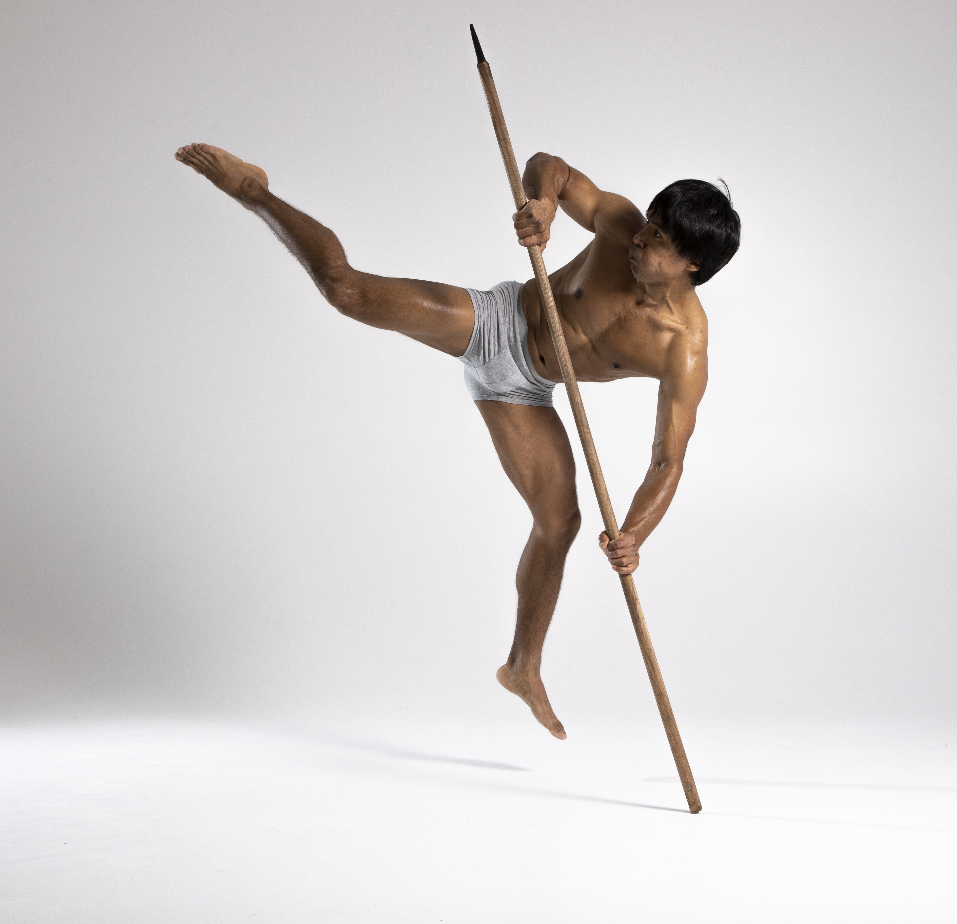 a man in a loincloth balances on one leg while holding a spear in a studio shot. he is barechested, showcasing his athletic build, and poses in a dynamic action pose reminiscent of a warrior or martial artist.