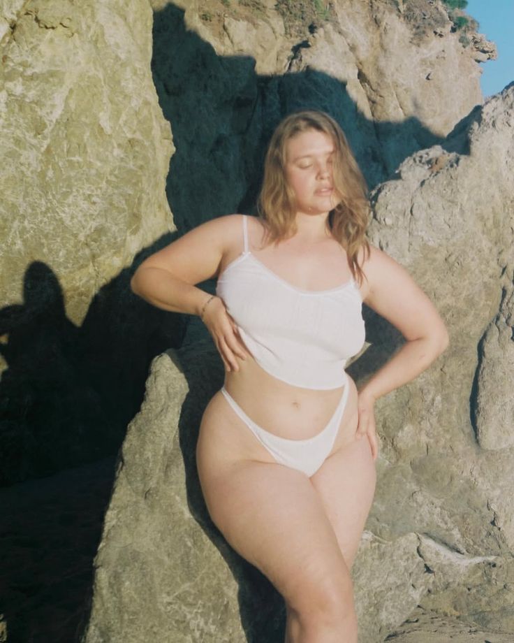 a woman with fair skin and blonde hair is posing on a beach. she is wearing a white twopiece swimsuit. she is standing in front of a large rock formation, with shadows cast on the rock. the sun is shining, creating a bright and natural light. her eyes are closed. she has a bodypositive pose, showcasing her curves.