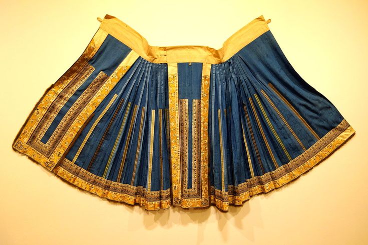 the image shows a traditional asian skirt, likely from the qing dynasty, displayed against a pale wall. the skirt is predominantly blue with gold embroidery and decorative borders. it is pleated and draped open, revealing its fanlike shape. the waistband is a light gold color. the skirt appears to be a cultural artifact, possibly displayed in a museum or gallery.