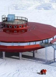 a red futuristic building with a circular domelike roof and windows sits on a snowy landscape in antarctica. the building appears to be a research station and has a large platform with stairs, railings, and walkways surrounding it. two figures in red and white clothes stand below the platform, looking towards the building. the landscape surrounding the building is covered in snow and ice.
