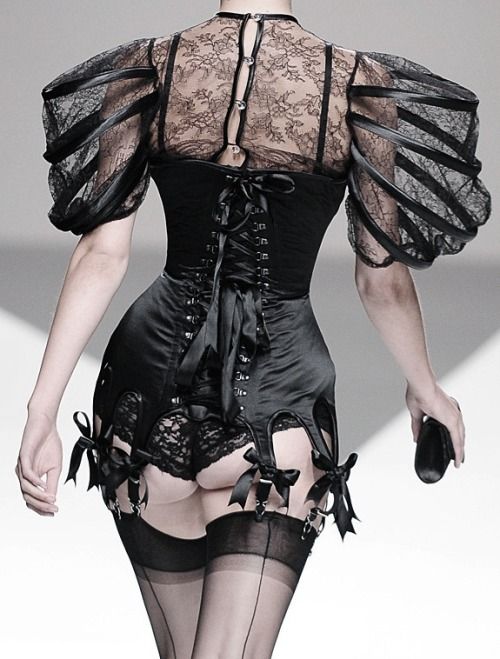 a woman in a black lace corset and stockings walks down a runway. her dress features an intricate lace design and a fitted bodice. she accessorizes with a black clutch and black high heels. the image is a fashion photograph, showcasing the beauty and elegance of the woman's outfit.