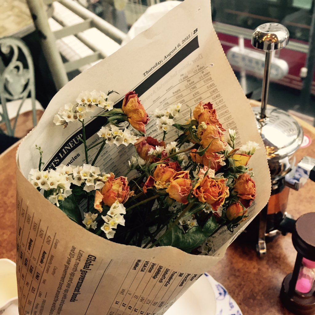 a bouquet of dried orange roses and white baby's breath wrapped in newspaper sits on a cafe table in front of a window. the table also has a tea set on it.