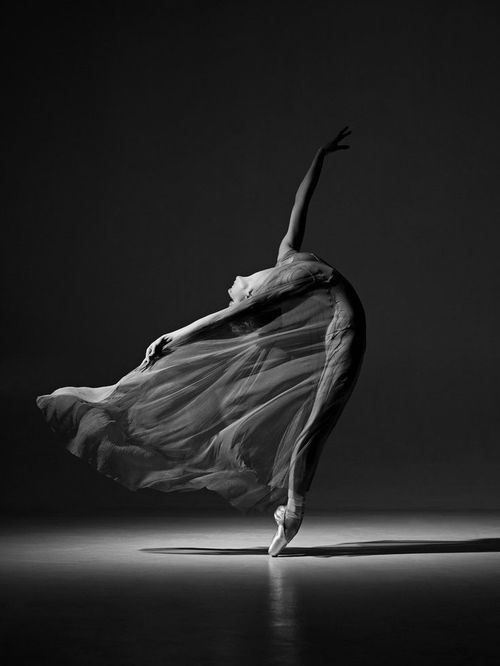 a black and white photograph of a ballerina in midair, her body contorted in a graceful pose. she is wearing a flowing dress that billows around her as she dances. the background is dark, highlighting her movement and the beauty of her form.
