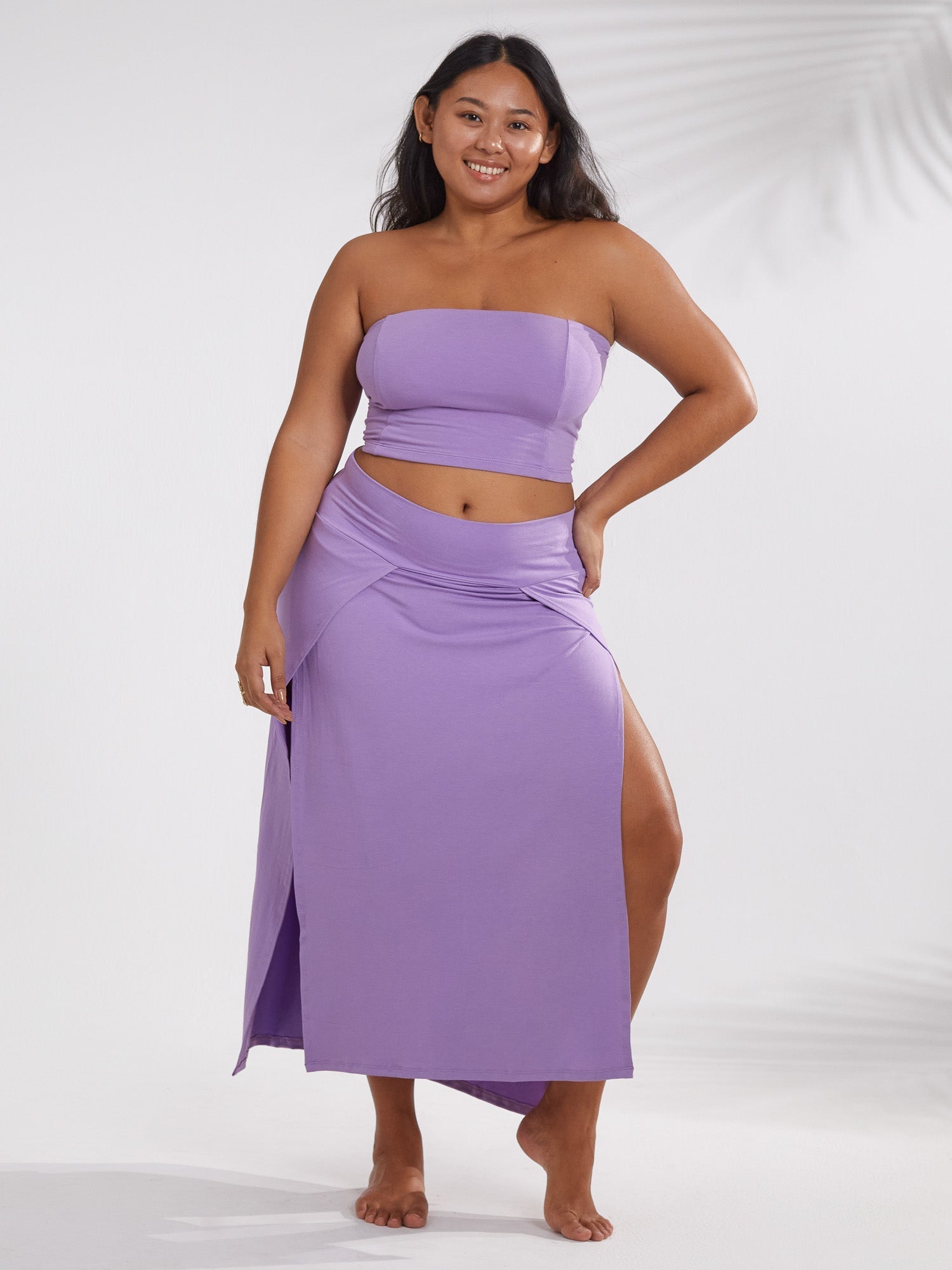 a fulllength studio shot showcases a plussize model wearing a stylish twopiece lavender outfit.  the ensemble consists of a strapless bandeau top and a matching maxi skirt featuring a thighhigh slit on one side. the model is standing with a relaxed posture, her dark hair falling around her shoulders. the background is a simple, lightcolored studio setting, allowing the outfit to be the focal point of the image. the fabric of the clothing appears smooth and soft. the overall style conveys a relaxed yet elegant summer aesthetic.
