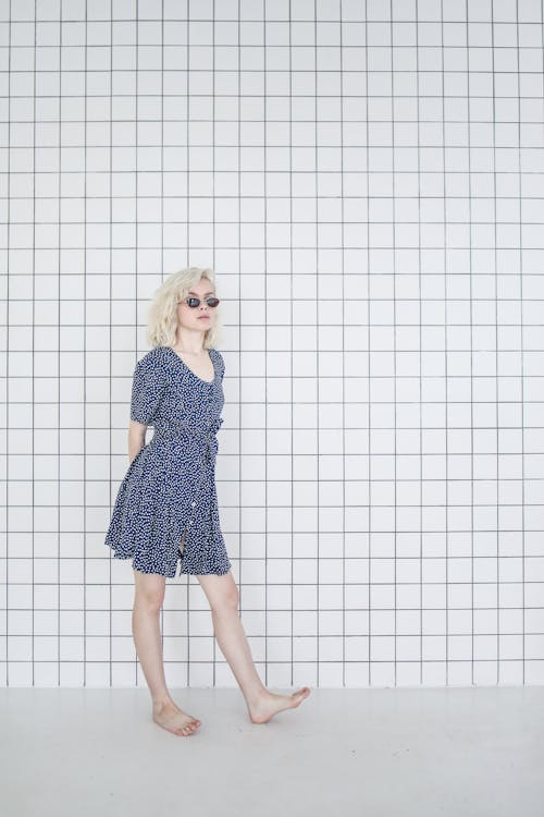 a woman with blonde hair and sunglasses wearing a blue polka dot dress stands against a tiled white wall.