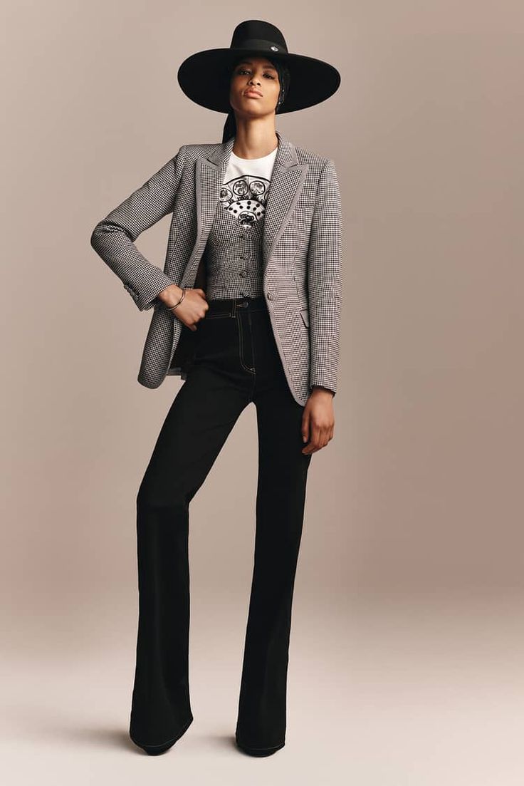 a female model is wearing a widebrimmed black hat, a white shirt, a grey checkered blazer, and black flare pants. the model is posing in a studio with a light brown background. she is looking at the camera with a confident expression.