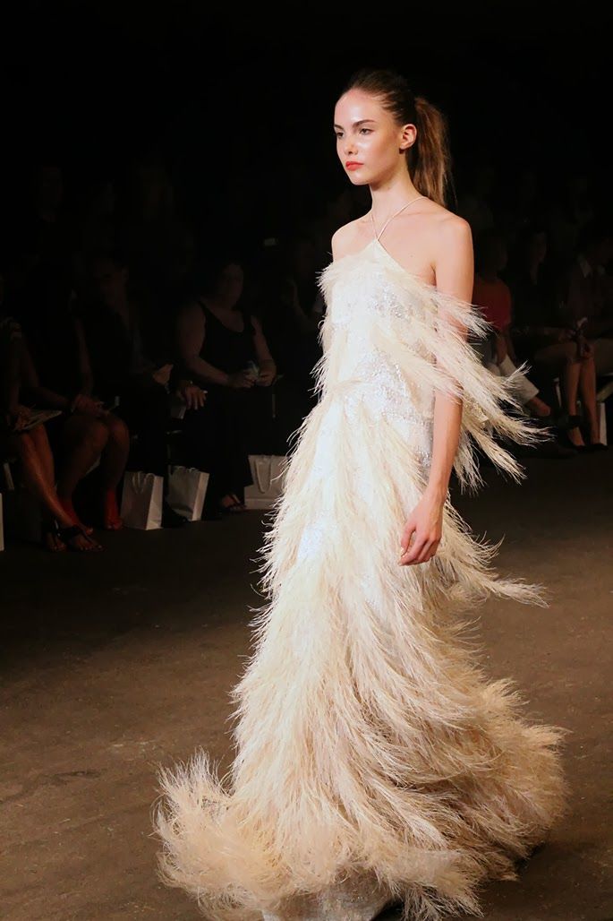 a model walks the runway in a long white feather dress. the dress is a glamorous and elegant design with a train trailing behind her. she is wearing simple makeup and has her hair pulled back.
