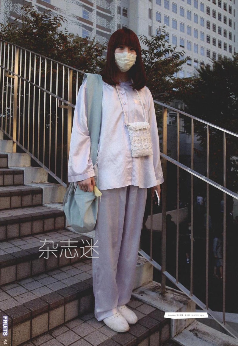 a young woman is standing on a set of stairs, posing for a street style photo. she is wearing a light purple satin pajamalike outfit, white sneakers, and a white face mask. she has a small white crocheted purse and a large sage green tote bag. the background is a city street with buildings and a railing. the photo has a vintage, slightly grainy aesthetic.