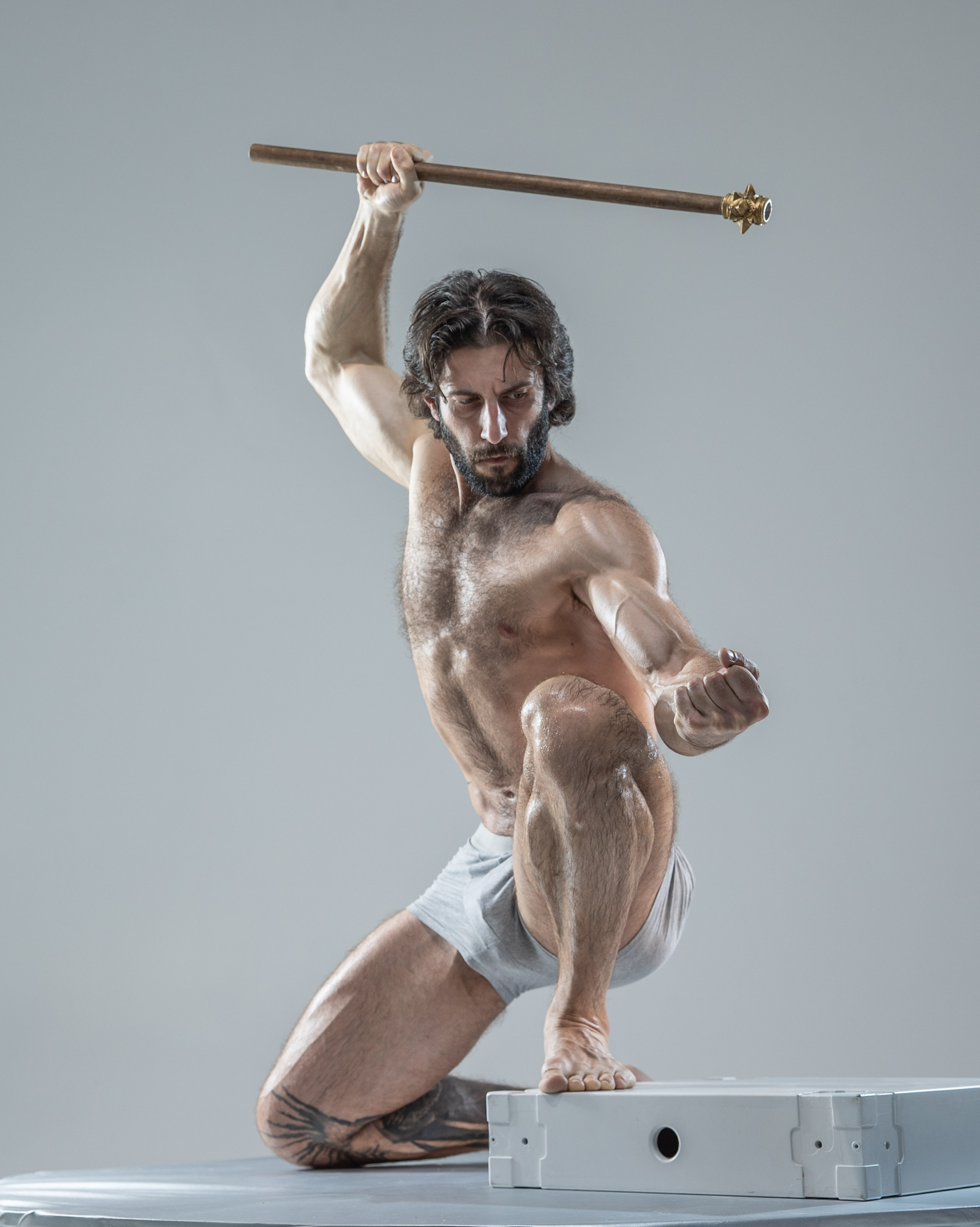 a muscular man with long hair and a beard, wearing white underwear, adopts an aggressive fighting stance against a light gray background. he holds a wooden staff with brass details, poised for attack. the image evokes strength, power, and the intensity of a warrior in action.