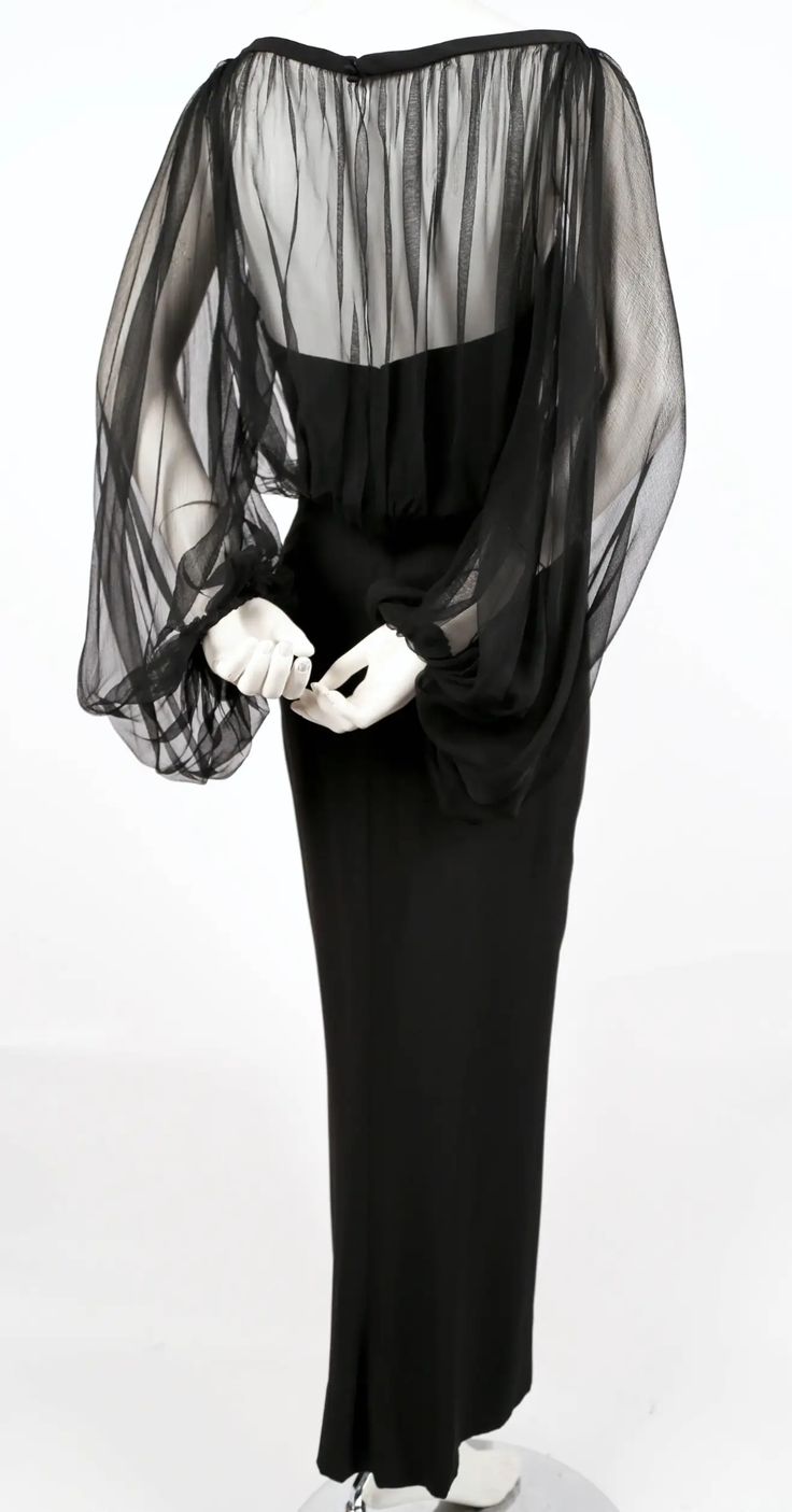 the image shows a black evening gown on a mannequin. the dress is floorlength with a fitted silhouette. it has a solid black bodice that transitions into a sheer black overlay on the upper portion of the dress, particularly around the shoulders and sleeves. the sleeves are voluminous and appear to be made of a thin, translucent fabric. the mannequin's hands are visible, positioned in front of the dress. the background is a plain white.