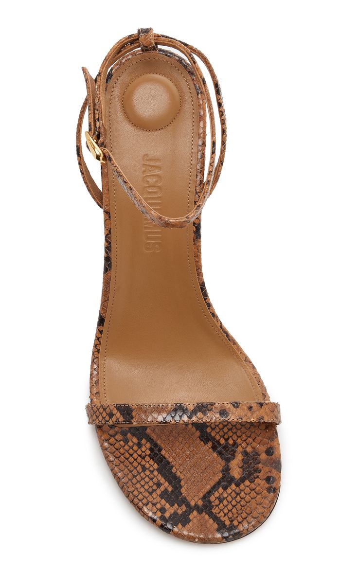 a topdown studio shot of a pair of jacquemus le bob sandals in brown snakeskin.  the sandals are highheeled with an ankle strap and are shown against a white background. the brand name "jacquemus" is visible on the insole.
