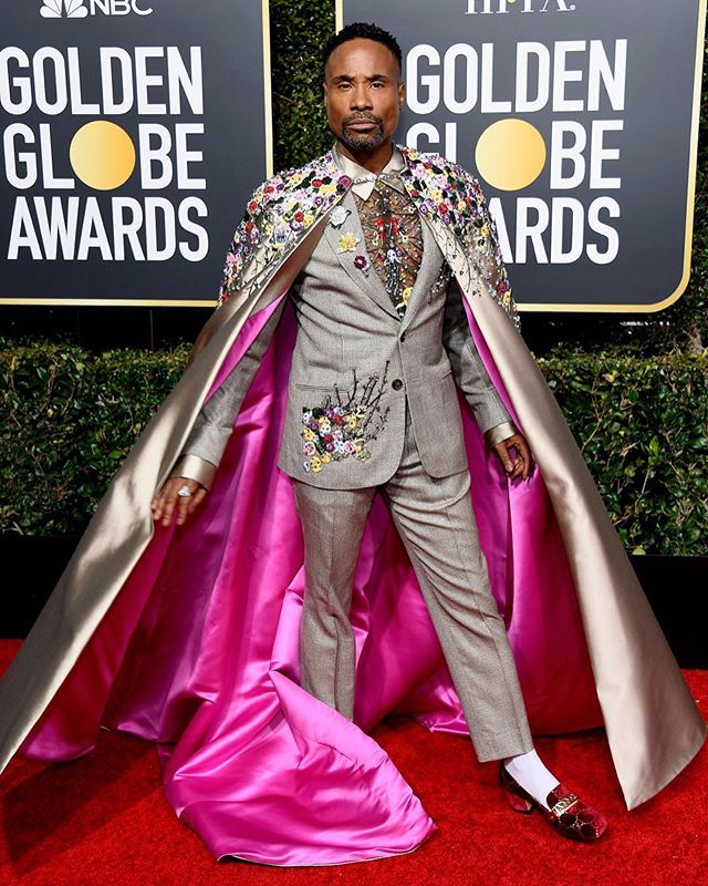 the image shows billy porter on the red carpet at the golden globe awards. he is wearing a gray suit with floral embroidery and a cape. the cape has a silver outer layer and a pink lining. he is also wearing unique shoes. the golden globe awards backdrop is visible behind him.