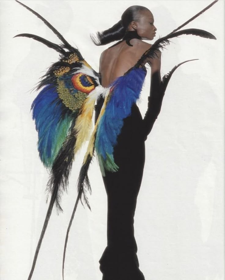 a fashion photograph showcases a woman with dark skin and sleek hair, standing with her back to the viewer. she is wearing a formfitting black gown and long black gloves, emphasizing her slender figure. the focal point is a pair of elaborate feather wings attached to her back, adorned with vibrant colors including blue, yellow, and green, reminiscent of peacock feathers. the overall impression is one of high fashion and artistic expression.