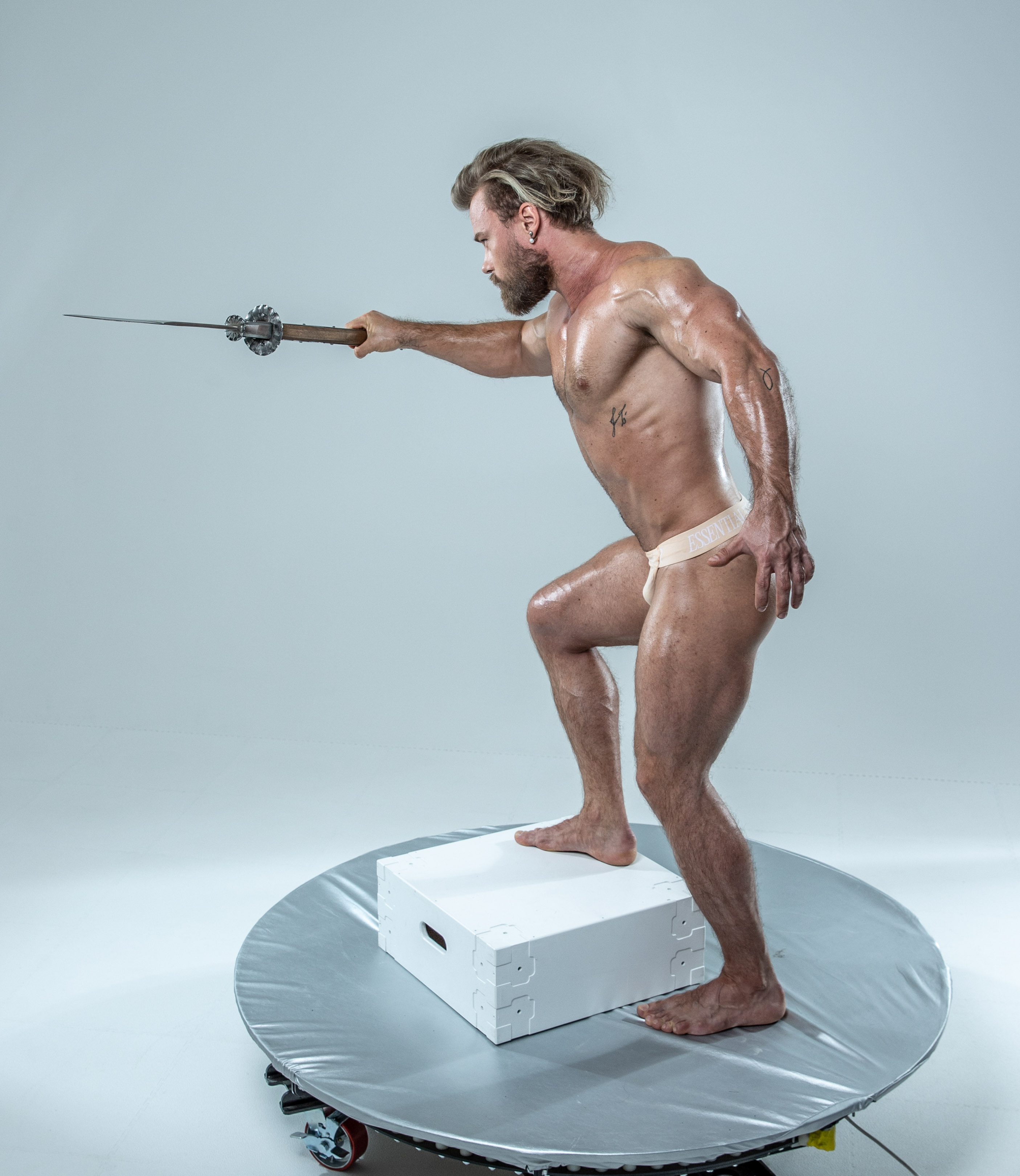 a muscular man with long blond hair is captured in a dynamic pose on a rotating platform against a white studio backdrop. he holds a medievalstyle sword out in front of him with his right hand while his left hand is clenched in a fist. he stands in a balanced stance with one foot on the platform and the other on a white box. the soft lighting highlights his physique and the overall image evokes a sense of power and athleticism.