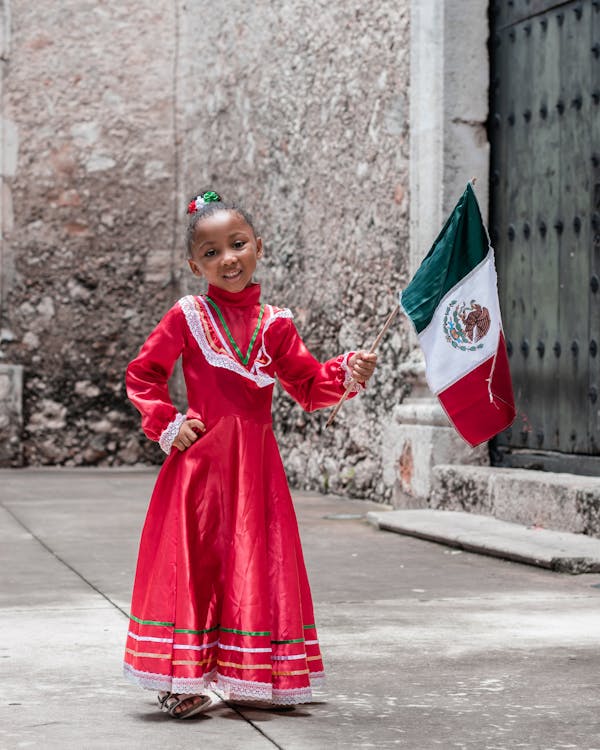 a young girl, dressed in a vibrant red traditional mexican dress, stands proudly holding a small mexican flag.  she is smiling happily and her dress features white lace and colorful stripes at the hem. the background is a rustic stone wall and street, creating a charming and patriotic scene.  the overall feeling of the image is one of joy and national pride.