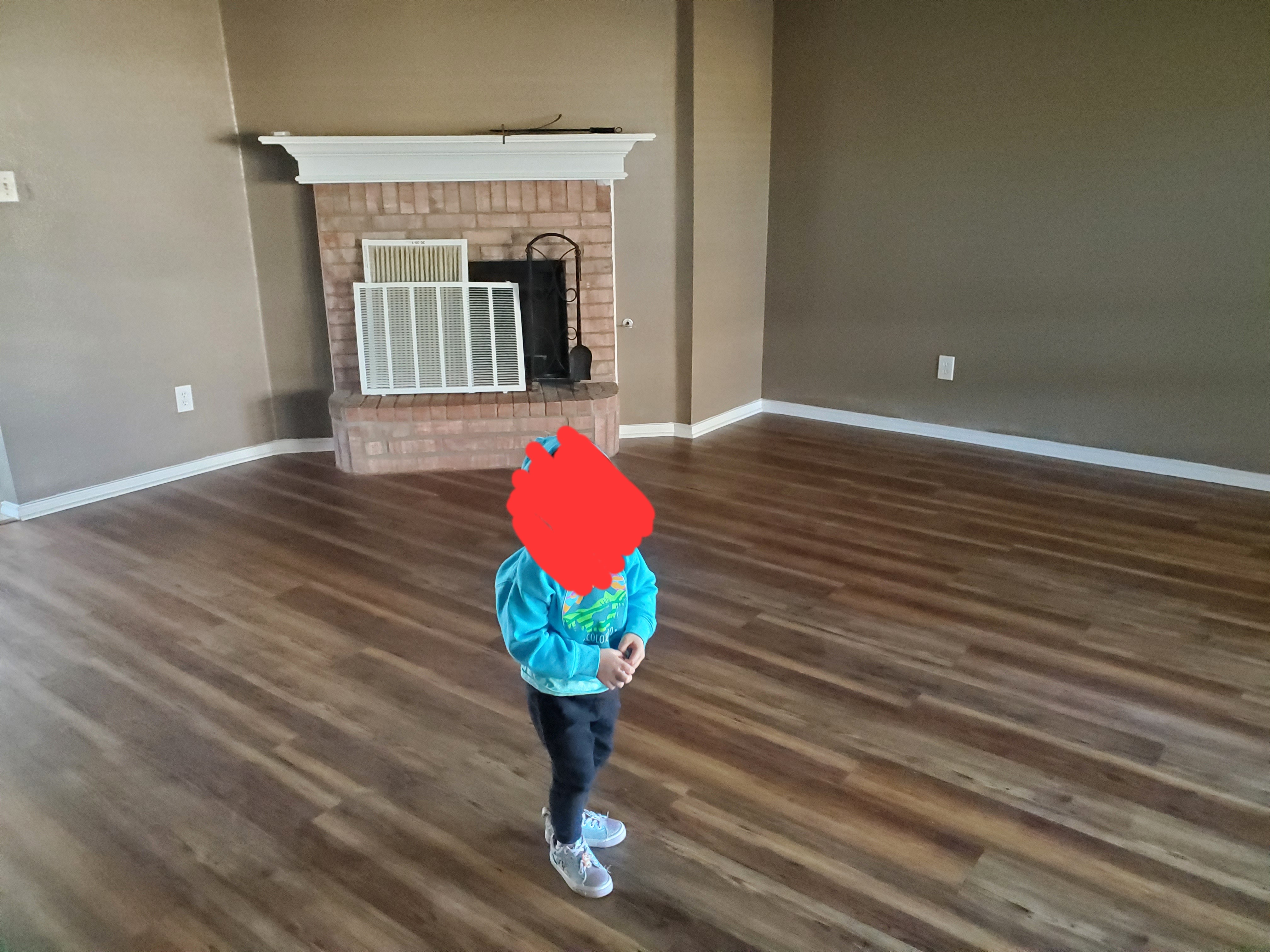 the image shows a living room with hardwood floors, a fireplace, and a child standing in the middle of the room.