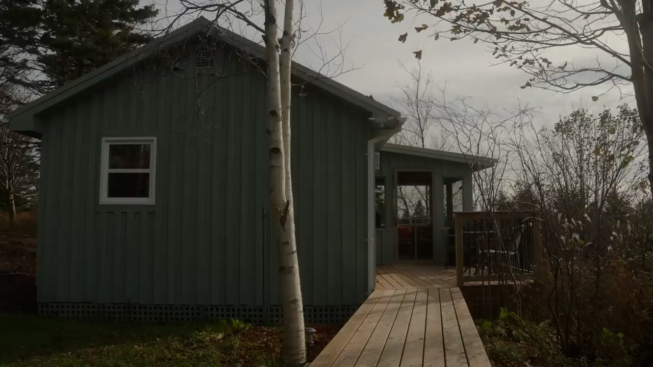 this is an image of a small green cabin in the woods. the cabin has a wooden deck and a porch. there are trees in the background.