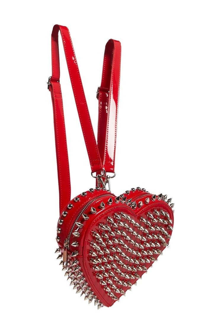 a red, heartshaped studded backpack is shown. the backpack is made of shiny red patent leather and covered with silvercolored studs. it has two red straps and two zippers at the top of each half of the heart. the background is plain white.
