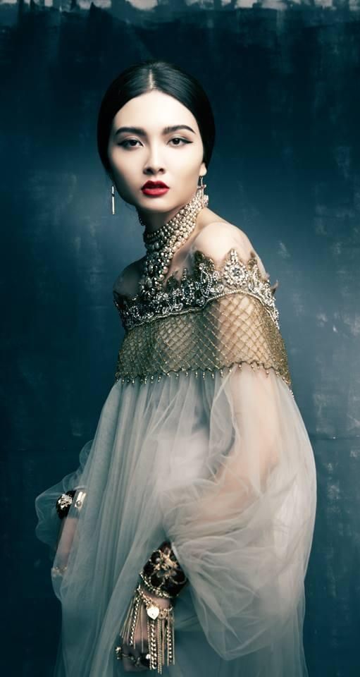 a stunning portrait of a woman adorned in a beautiful gown and elegant jewelry. she has a captivating gaze, accentuated by her striking makeup and asian features. the gown showcases intricate gold detailing, adding to the overall opulence and fine art aesthetic.