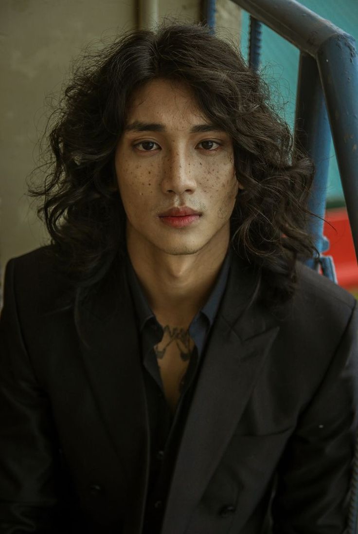 closeup portrait of a young man with long, curly dark hair and freckles. he's wearing a darkcolored suit jacket and a collared shirt. his expression is serious and neutral. he appears to have a small tattoo on his chest. the background is slightly blurred, showing a wall and part of a staircase. the overall mood is moody and atmospheric.