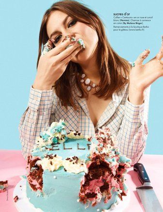 a young woman is eating a large, messy cake with a bright blue frosting. the cake is surrounded by sprinkles and has a small red heart on the side. the woman is smiling and appears to be enjoying the cake. she is wearing a plaid shirt and a necklace and has a big, playful smile. the image is bright and cheerful and captures the fun and joy of celebrating with cake.