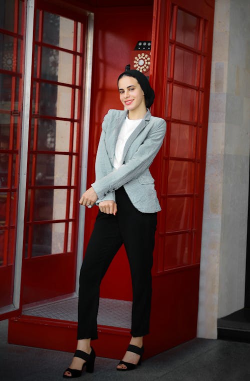 a stylish young woman wearing a hijab stands confidently in front of a classic red london telephone booth. she is dressed in a light blue blazer, black pants, and black heels. her outfit is modern and elegant. the background is blurred but shows the red booth and a hint of a city street. the image is welllit and focuses on the woman's poise and fashion sense.